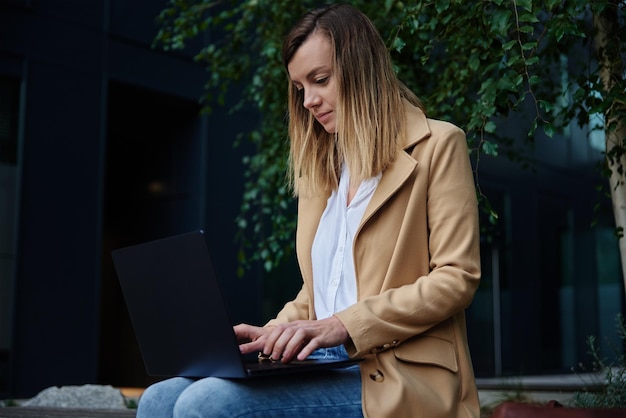 Frau, die aus der Ferne arbeitet, sitzt auf einer Bank in der Stadtstraße und arbeitet online mit Laptop-Freiberuflerinnen