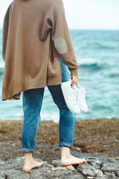 Frau, die barfuß am Meer spaziert und Händchen hält, weiße Schuhe Sommerurlaub