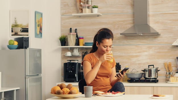 Frau, die beim Frühstück telefoniert und frischen Orangensaft mit einer gesunden Bio-Mahlzeit und einer Tasse Kaffee trinkt, wobei sie moderne Technologie allein in der Küche verwendet, bevor sie zur Arbeit geht.