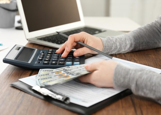 Frau, die Berechnungen anstellt, während sie Dollarscheine über Steuerformular auf dem Holztisch hält, hautnah
