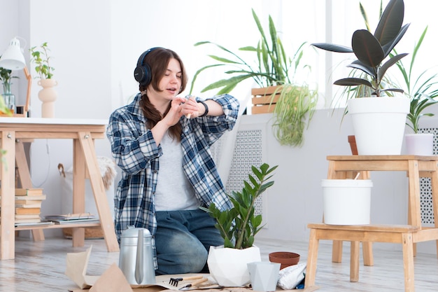 Frau, die die Pflanze in einen neuen Topf verlegt und zu Hause Musik mit drahtlosen Kopfhörern und einer intelligenten Uhr hört