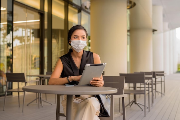 Frau, die draußen im Café-Restaurant sitzt, soziale Distanzierung und Gesichtsmaske trägt, um sich vor Covid 19 zu schützen, während sie die horizontale Aufnahme von Telefon und digitaler Tablette verwendet.