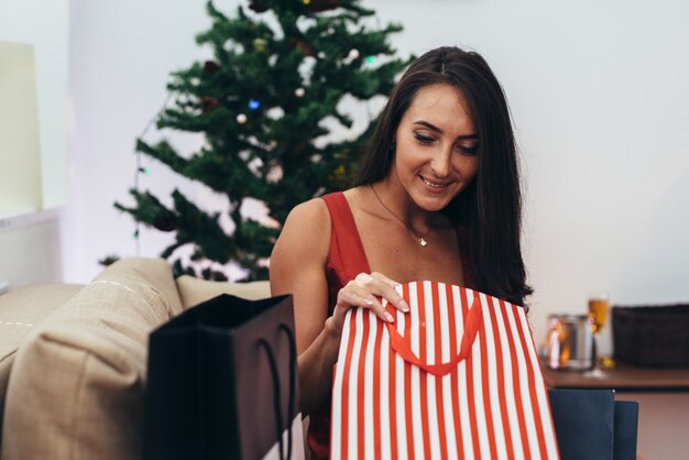 Frau, die durch den Weihnachtsbaum stationiert und ein Geschenk aus Einkaufstasche heraus nimmt