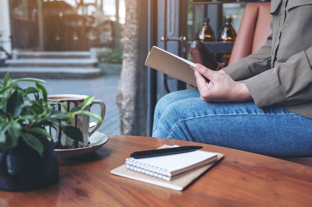 Frau, die ein Buch mit Notizbüchern und Kaffeetasse auf Holztisch im Café öffnet