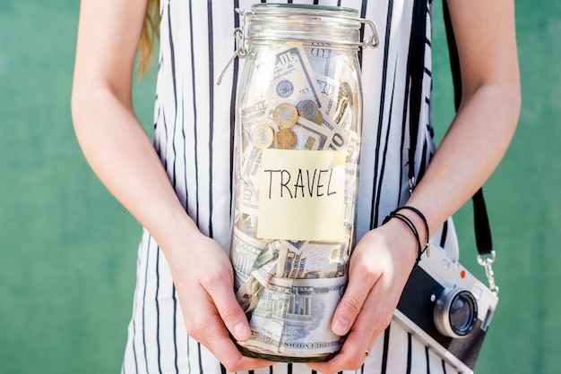 Frau, die ein Glas voller Geld für das Reisen hält, das mit Fotokamera auf dem grünen Hintergrund steht. Geld sparen für Sommerferienkonzept