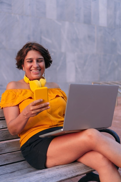 Frau, die ein Handy und einen Laptop benutzt, während sie auf einer Bank im Park sitzt