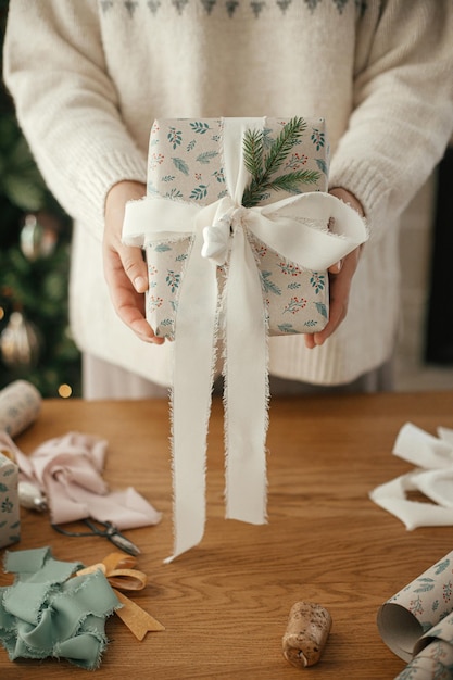 Frau, die ein stilvoll geschmücktes Weihnachtsgeschenk auf dem Hintergrund eines Tisches mit festlichen Dekorationen in einem modernen skandinavischen Zimmer hält.