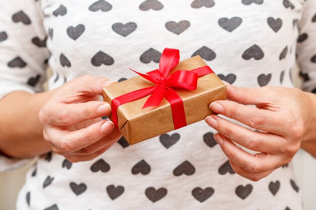 Frau, die eine Geschenkbox mit einem roten Band in ihren Händen hält. Geringe Schärfentiefe, selektiver Fokus auf die Box. Konzept, am Valentinstag oder Geburtstag ein Geschenk zu geben.