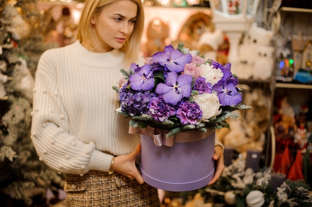 Frau, die eine riesige Schachtel mit lila und violetten Blumen hält, die mit Tannenzweigen verziert sind