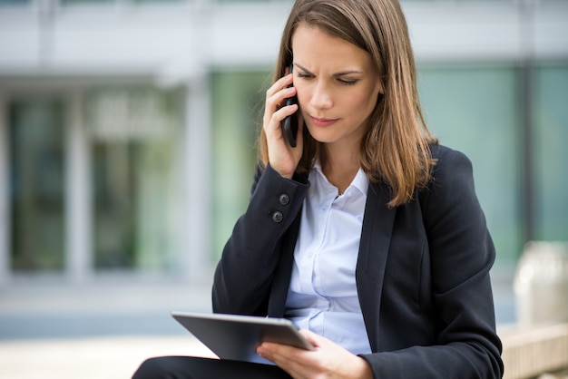 Frau, die eine Tablette bei der Unterhaltung an ihrem Mobilhandy vor ihrem Büro verwendet