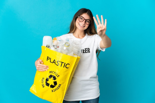 Frau, die eine Tasche voller Flaschen hält, um lokalisiert auf blauer Wand glücklich zu recyceln und vier mit den Fingern zu zählen