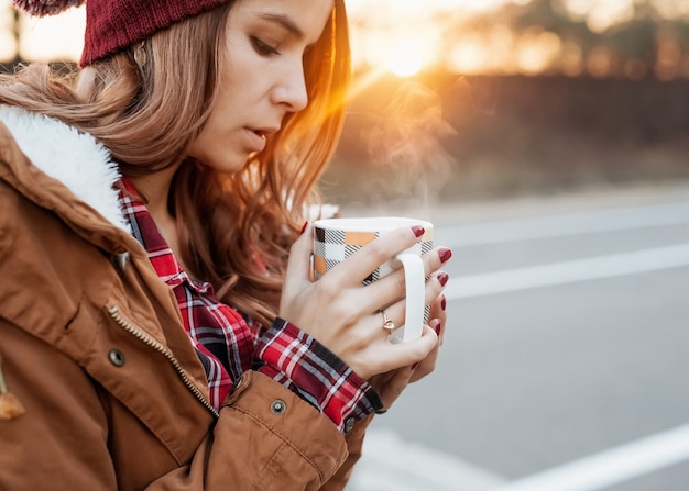 Frau, die eine tasse heißes getränk mit dämpfen im wintersonnenuntergangslicht hält.