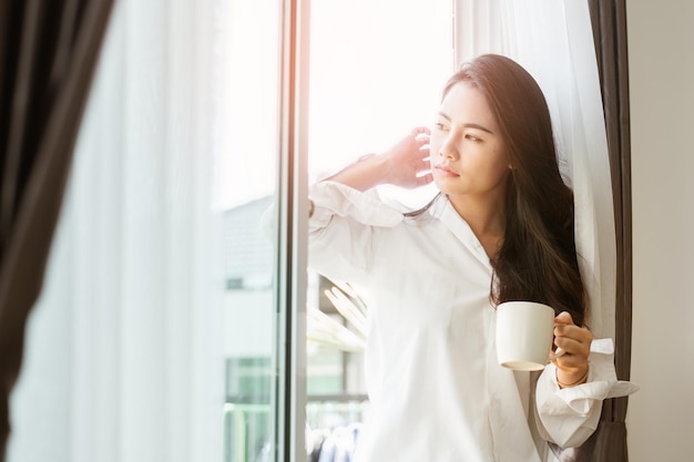 Frau, die eine Tasse Kaffee in ihren Händen hältFrau trinkt Kaffee zu Hause mit Sonnenaufgang am Fenster