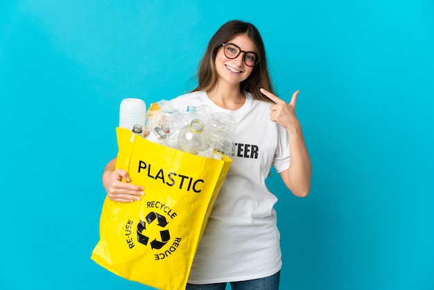 Frau, die einen Beutel voller Flaschen hält, um lokalisiert auf der blauen Wand zu recyceln, die eine Daumen hoch Geste gibt