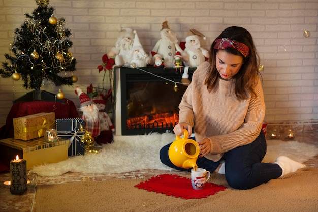 Frau, die einen heißen Kaffee nahe bei dem Kamin mit Weihnachtsatmosphäre gießt.