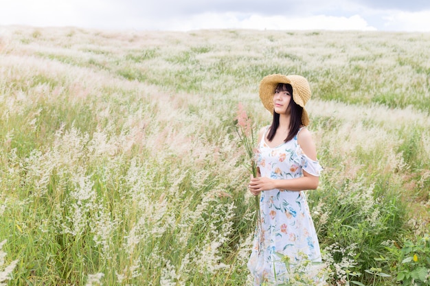 Frau, die einen Hut trägt, ein weißes Kleid trägt und mitten im Gras mit schönen weißen Blumen mit einer entspannten und glücklichen Stimmung steht.