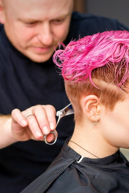 Frau, die einen neuen Haarschnitt hat. Ein männlicher Friseur schneidet in einem Friseursalon gefärbte rosa kurze Haare mit einer Schere.