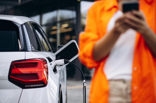 Frau, die Elektroauto an der Ladestation auflädt und Handy benutzt