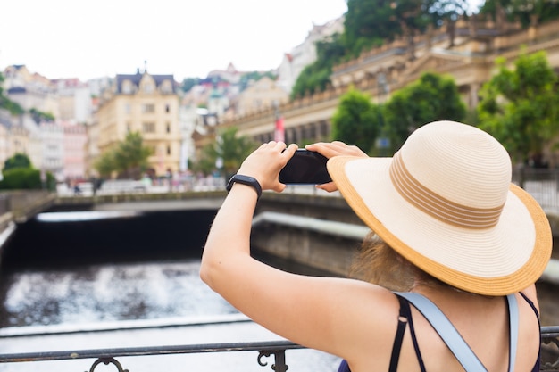 Frau, die Fotos mit stilvoller Sommerreisenderfrau des Smartphones mit Hut mit Kamera draußen macht