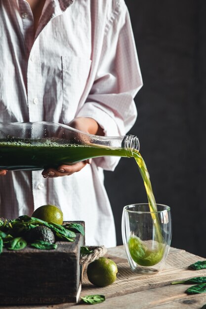 Frau, die frisch gemischten grünen und weißen Smoothie im Glasglas mit Bestandteilen herum auf einem rustikalen Holztisch gießt