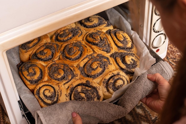 Frau, die gebackene Brötchen mit Mohn aus dem Ofen nimmt Fertiger frischer Kuchen mit Mohn, der zu Hause gebacken wird