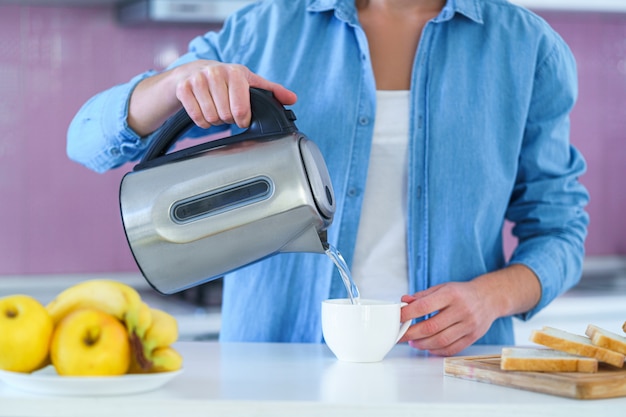 Frau, die gekochtes Wasser in eine Tasse von einem elektrischen Kessel gießt, um heißen Tee zu Hause in der Küche zu brauen