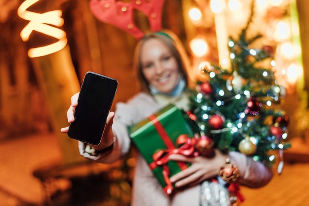 Frau, die Geschenke, Weihnachtsbaum und Telefon mit einem schwarzen Bildschirm hält