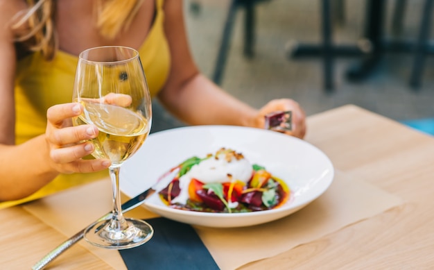 Frau, die gesunden Salat mit Burrata-Käse, Rucola-Salat und Tomaten isst und Weißwein in einem Glas hält