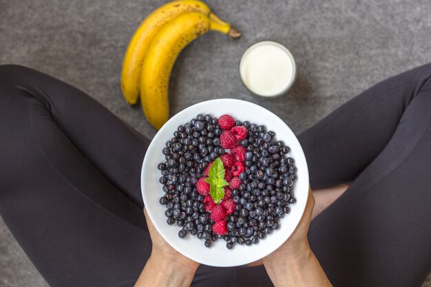 Frau, die gesundes Lebensmittel isst. Yong-Mädchen in der Sportkleidung, die einen Teller von frischem berris hält. Bananen und Milchprodukte in der