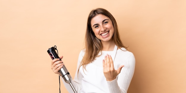 Frau, die Handmixer über isolierte Wand verwendet, die einlädt, mit Hand zu kommen. Schön, dass du gekommen bist