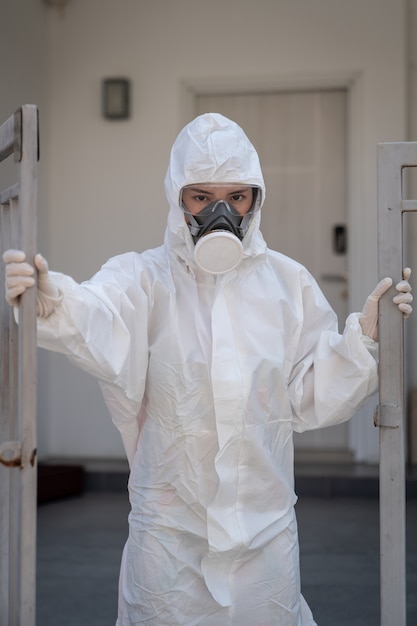 Frau, die Handschuhe mit chemischem Schutzanzug und Maske der biologischen Gefahr mit unglücklichem Gesicht trägt. Ein Eisentor öffnen.