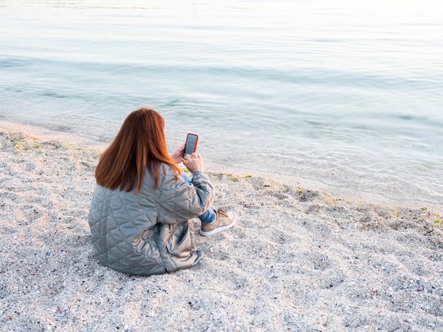 Frau, die Handy benutzt, während sie auf Strandsand sitzt