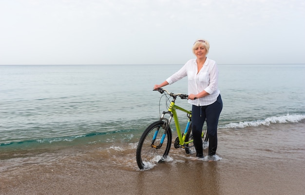 Frau, die ihr Fahrrad durch das flache Meerwasser am Rand des Strandes an einem wolkigen bewölkten Tag dreht