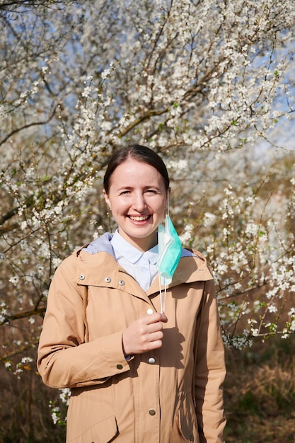 Frau, die im Frühling an einer saisonalen Allergie leidet, posiert im blühenden Garten im Frühjahr, entfernt eine medizinische Maske vor blühenden Bäumen und lächelt nach der Behandlung.