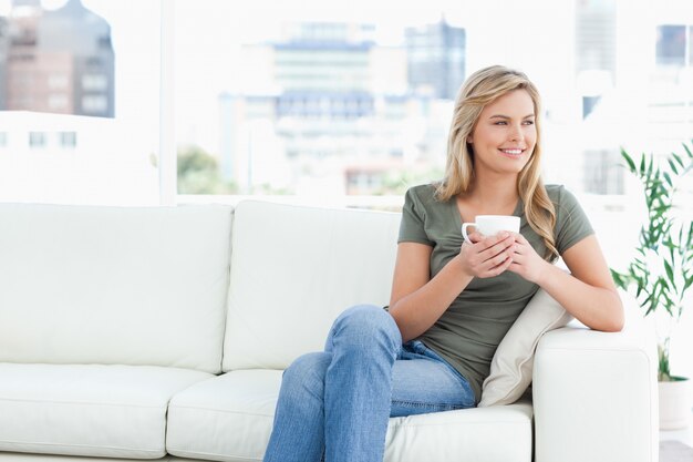 Frau, die in der Hand auf der Couch, mit Becher sitzt, seitlich lächelt und schaut