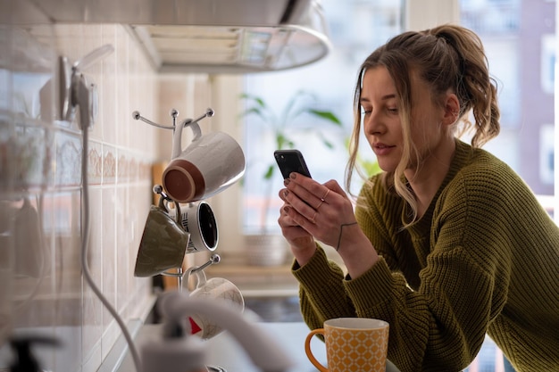 Frau, die in der Küche steht und eine Tasse Tee trinkt, während sie das Smartphone benutzt