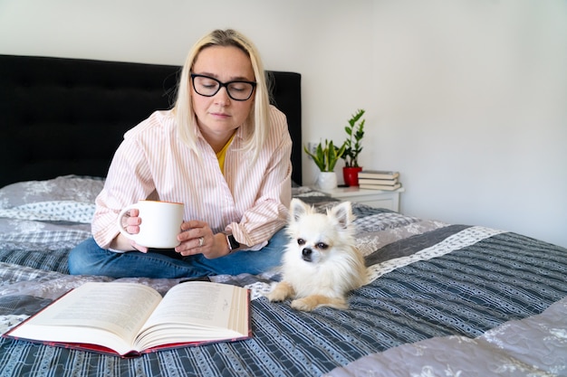 Frau, die in einem gemütlichen Bett mit ihrem Hund liegt und ein Buch liest
