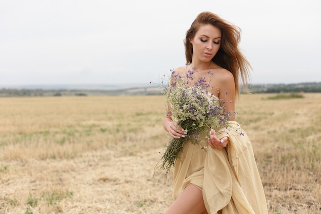 Frau, die in goldenes getrocknetes Grasfeld geht. Natürliche Porträtschönheit. Schönes Mädchen auf einem Weizengebiet. Junge Frau in einem beigefarbenen Kleid mit einem Strauß Wildblumen.