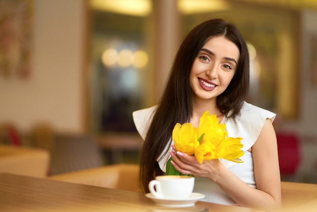 Frau, die Kaffee trinkt und Telefon benutzt. Frühlingsstil. Brünette Frau Im schönen Frühling. Schöne junge Frau mit Frühlingstulpenblumenstrauß am Café. schöne Frühlingsstimmung