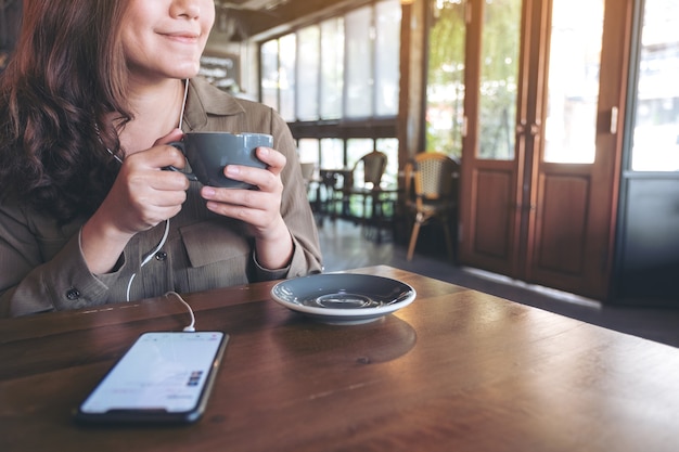 Frau, die Kaffee trinkt, während sie Musik mit Handy und Kopfhörer im Café hört