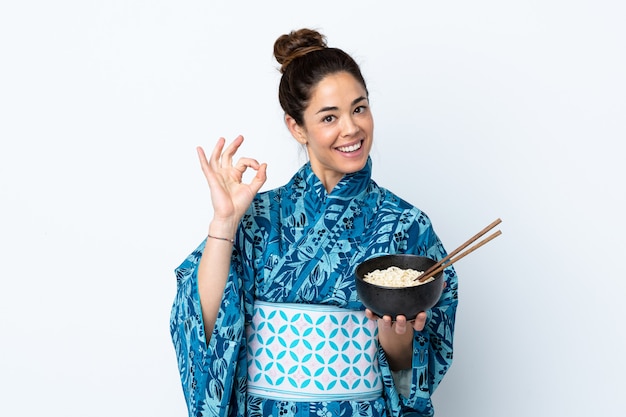 Frau, die Kimono über isoliertem Weiß trägt, das ok Zeichen mit den Fingern zeigt, während eine Schüssel Nudeln mit Stäbchen hält