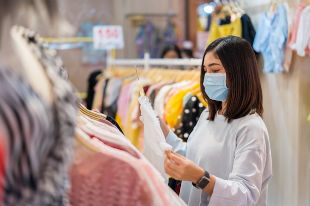 Frau, die Kleidung am Einkaufszentrum wählt und medizinische Maske zur Verhinderung von Coronavirus trägt