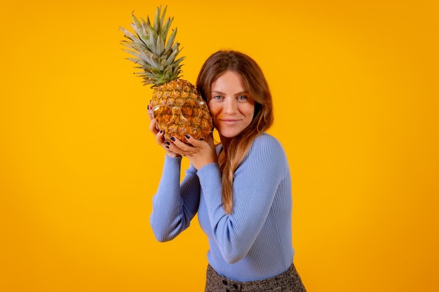 Frau, die mit einer Ananas in der Sonnenbrille in einem Studio auf einem gesunden Sommer des gelben Hintergrundes lächelt