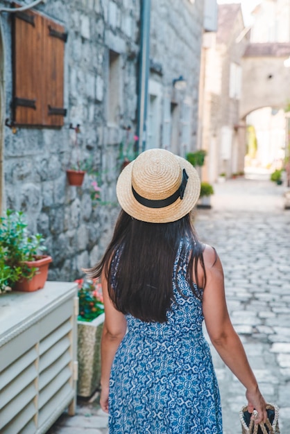 Frau, die mit Hut und blauem Kleid durch kleine Perast-Straßen geht