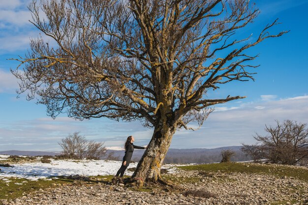 Frau, die mit ihrer Hand steht, die auf einem großen blattlosen Baum mitten in einer Winterlandschaft mit Schnee und Schmutz ruht. Umweltschutz