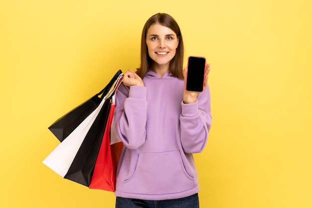 Frau, die mit zufriedenem Blick steht und Einkaufstaschen und Smartphone mit leerem Bildschirm für Werbung zeigt