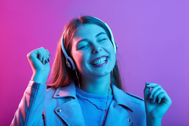 Frau, die Musik über Kopfhörer hört, hält Augen geschlossen und glücklich lächelnd, hält Fäuste isoliert über rosa Neonraum heben