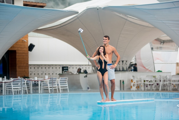 Frau, die nahe dem swimmingpool macht selfie foto mit dem einbeinstativ und mann steht hinter ihr aufwirft