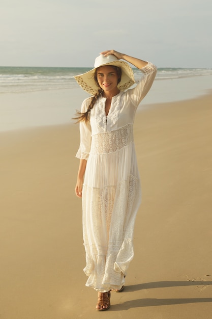 Frau, die schönes weißes Kleid trägt, geht am Strand während des Sonnenuntergangs