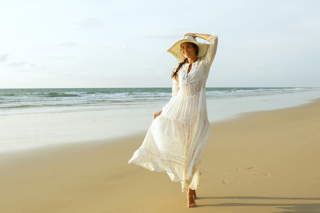 Frau, die schönes weißes Kleid trägt, geht am Strand während des Sonnenuntergangs